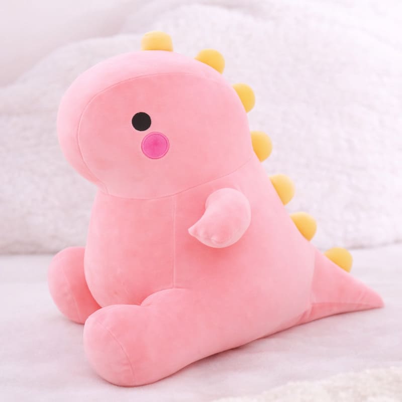 Octoplush Kawaii collection pink dinosaur plushie, a soft and squishable pastel pink dino stuffed animal sitting on a white bed. This cute Octoplush.store toy features yellow back spikes and bright pink rosy cheeks, making it an essential piece for aesthetic room decor and the perfect gift for girls.