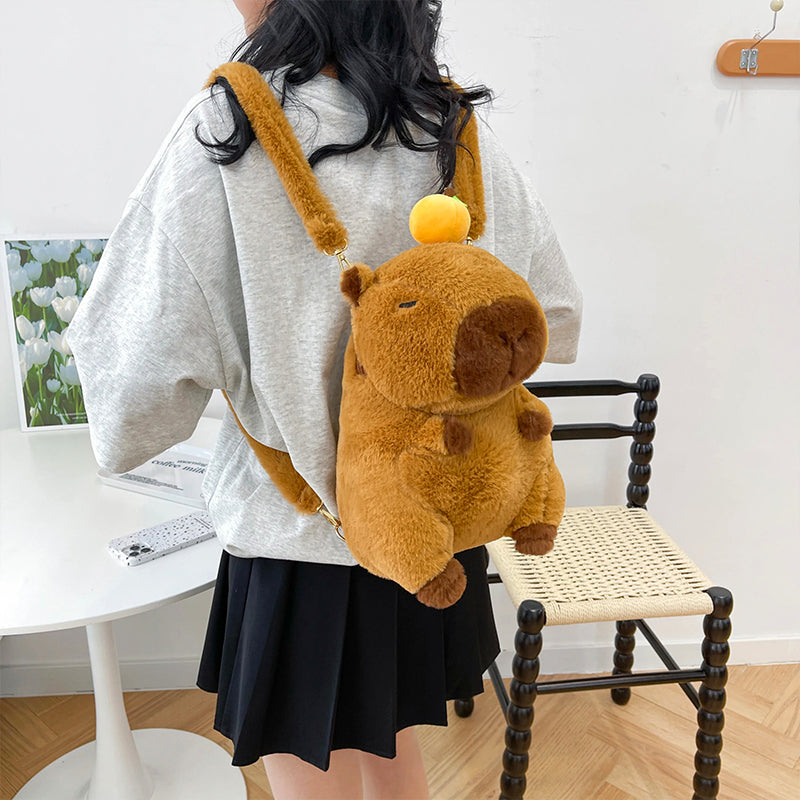 Fluffy capybara backpack with orange hat worn by girl in grey tee and black skirt, indoor lifestyle setup.