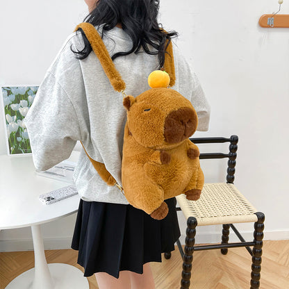 Fluffy capybara backpack with orange hat worn by girl in grey tee and black skirt, indoor lifestyle setup.