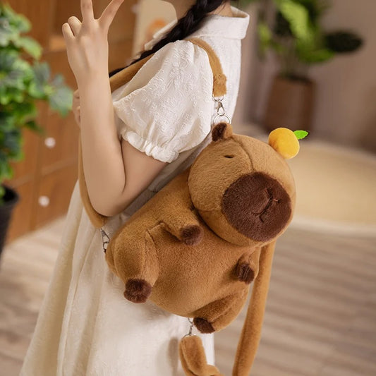 Capybara plush backpack with sleepy face and fruit hat, worn crossbody by girl in white dress flashing peace sign.