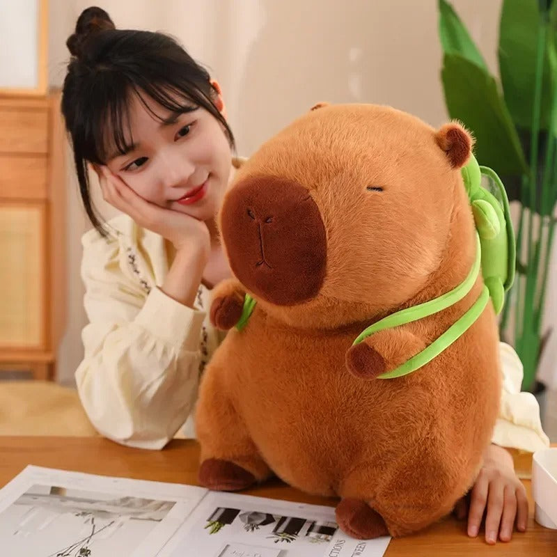 Young woman gazes affectionately at a giant capybara plush toy with a green turtle backpack, seated together at a wooden desk in a cozy room.