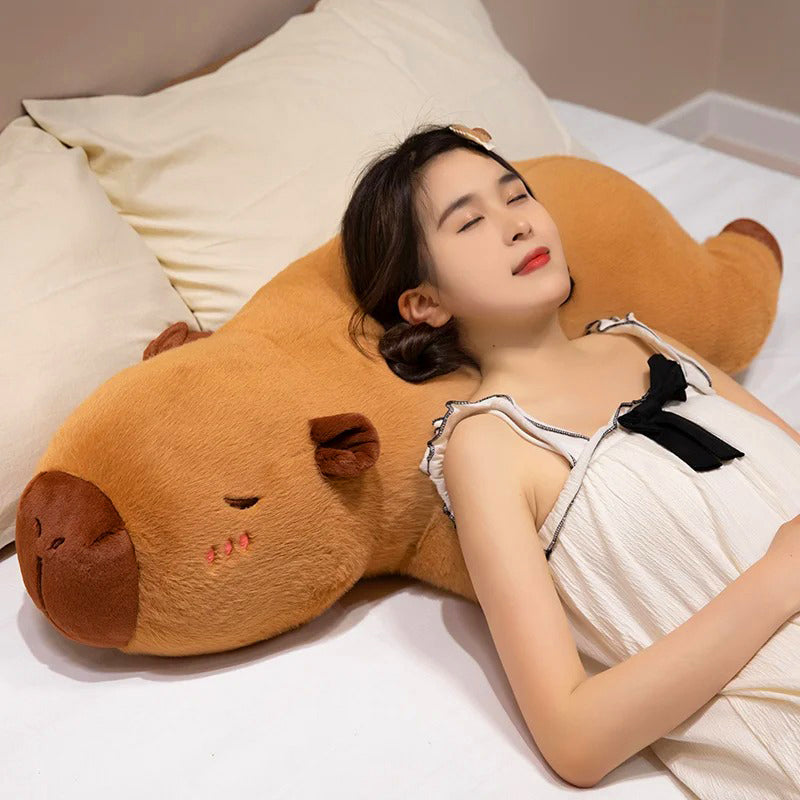 Woman resting her head on an oversized capybara stuffed animal pillow while relaxing in bed.