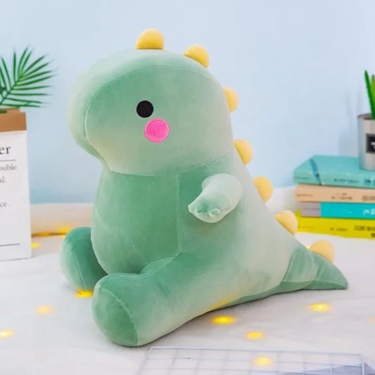 A soft and squishable green dinosaur plushie from the Kawaii collection, featuring rosy pink cheeks and yellow spikes, sitting on a white surface with warm fairy lights.