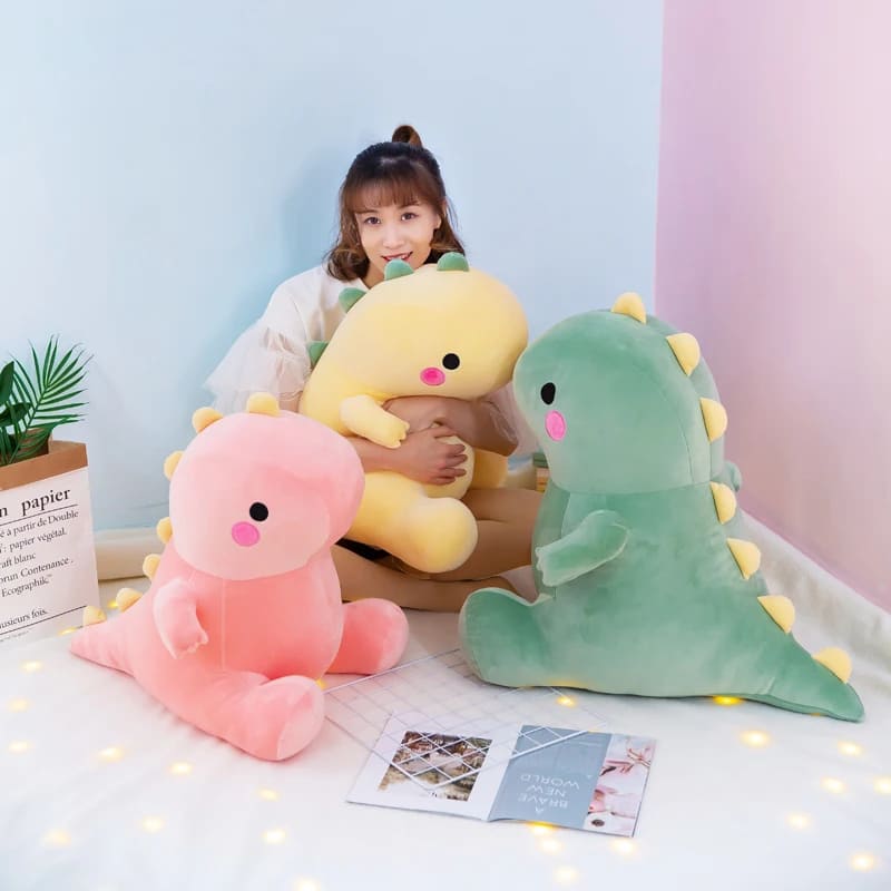 Kawaii collection dinosaur plushies in pink, yellow, and green. These soft and squishable stuffed animals are shown in different sizes, featuring cute rosy cheeks and yellow spikes. A girl holds a medium yellow dino, while a large green dinosaur and a small pink plushie sit on a white surface, perfect for pastel bedroom decor.