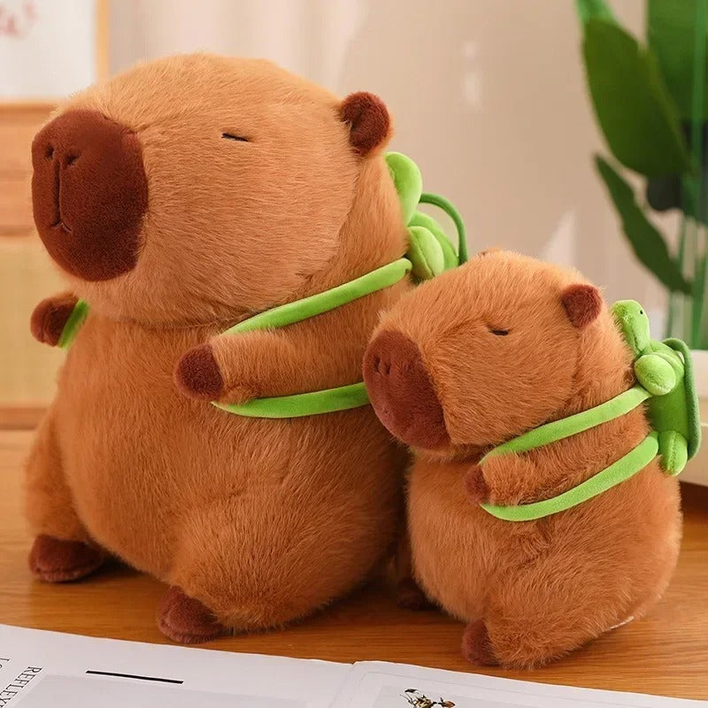 Side angle of two capybara plushies with matching green turtle backpacks, one medium-sized and one small, posed together on a desk to display their size comparison.